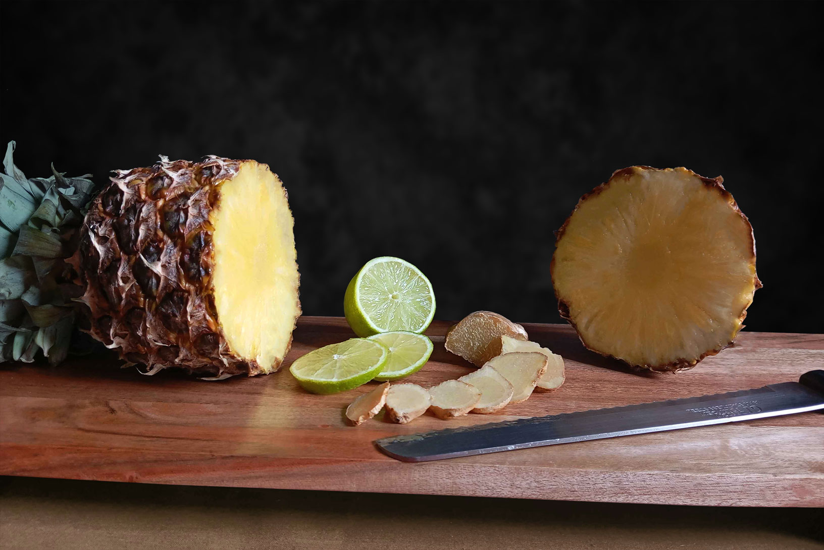 Photo-présentation-2-web Une photographie professionnelle d'un ananas coupé en deux sur une planche en bois. Un citron vert et du gingembre ornent le tout.